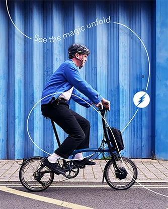 A woman sits on stairs with a folded bicycle and a small backpack beside her.